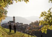 Ein Mann und eine Frau wandern einen Wiesenweg entlang und blicken auf Völs am Schlern