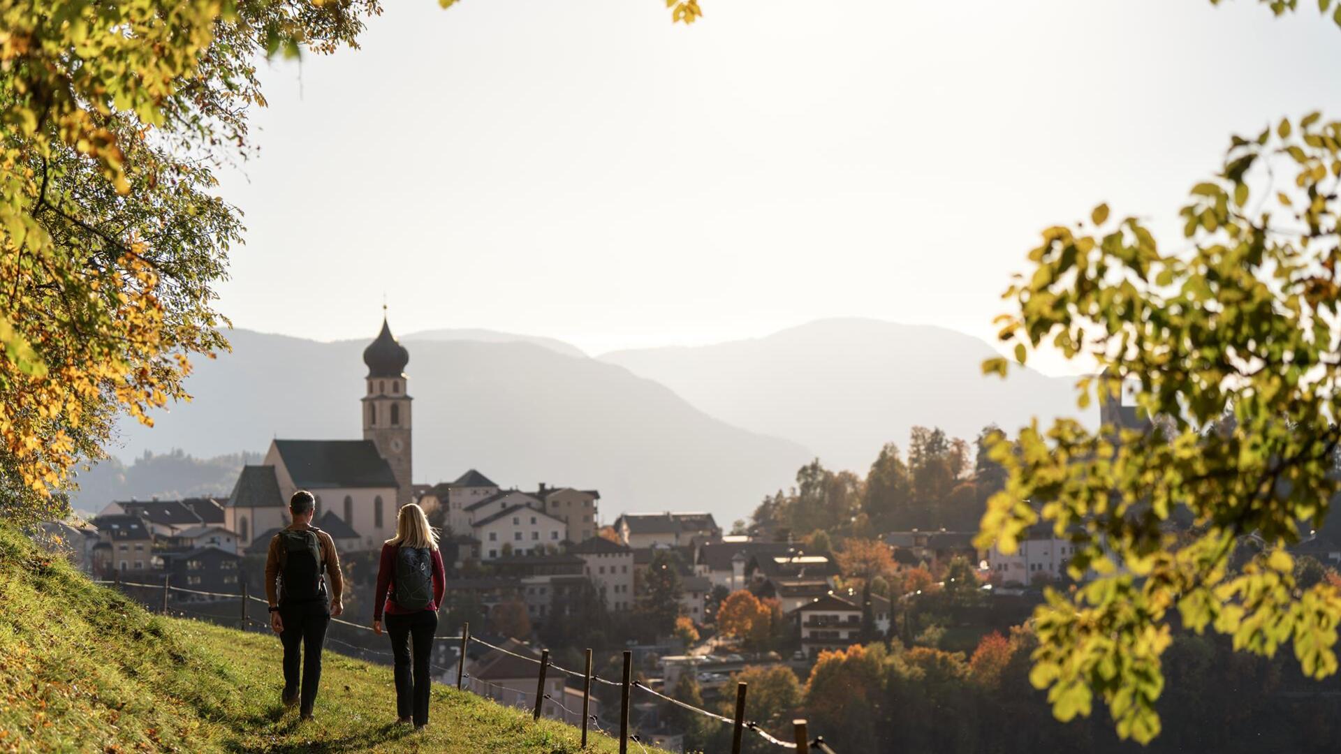 A man and a woman walk along a meadow path and look toward Völs am Schlern