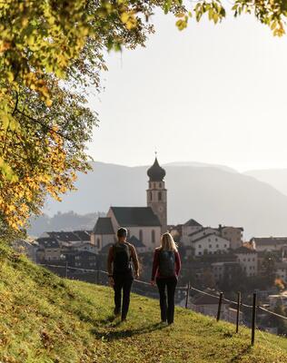 Ein Mann und eine Frau wandern einen Wiesenweg entlang und blicken auf Völs am Schlern