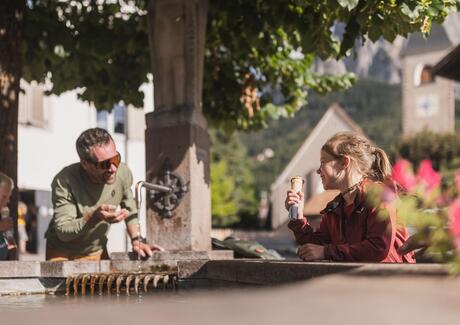 Ein Mann trinkt am Brunnen Wasser, und ein Mädchen isst daneben ein Eis