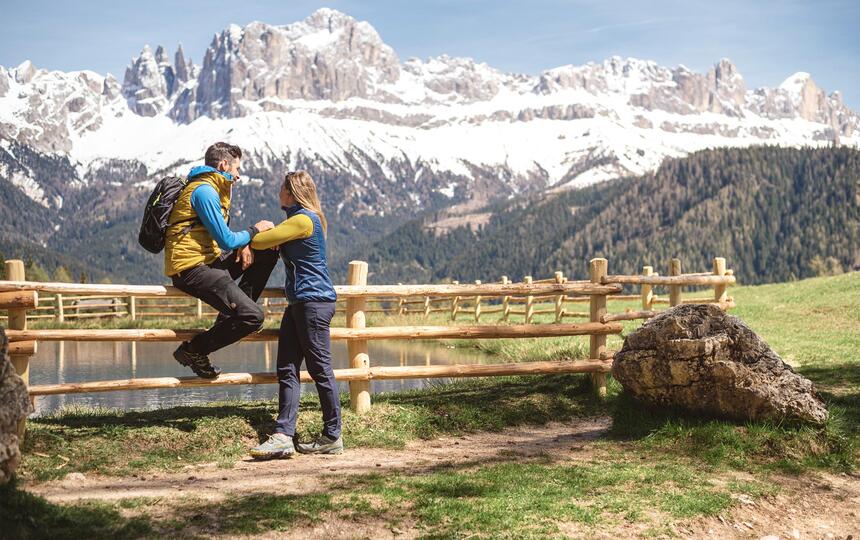 Un uomo è seduto su una staccionata vicino a un lago, una donna è in piedi accanto a lui e entrambi guardano le Dolomiti