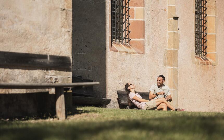 Un uomo e una donna sono seduti su una panchina di legno; sullo sfondo si vede la Chiesa di San Valentino