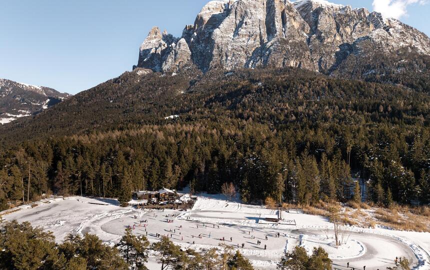 Luftbild des zugefrorenen Völser Weihers; zahlreiche Personen laufen darauf Schlittschuh