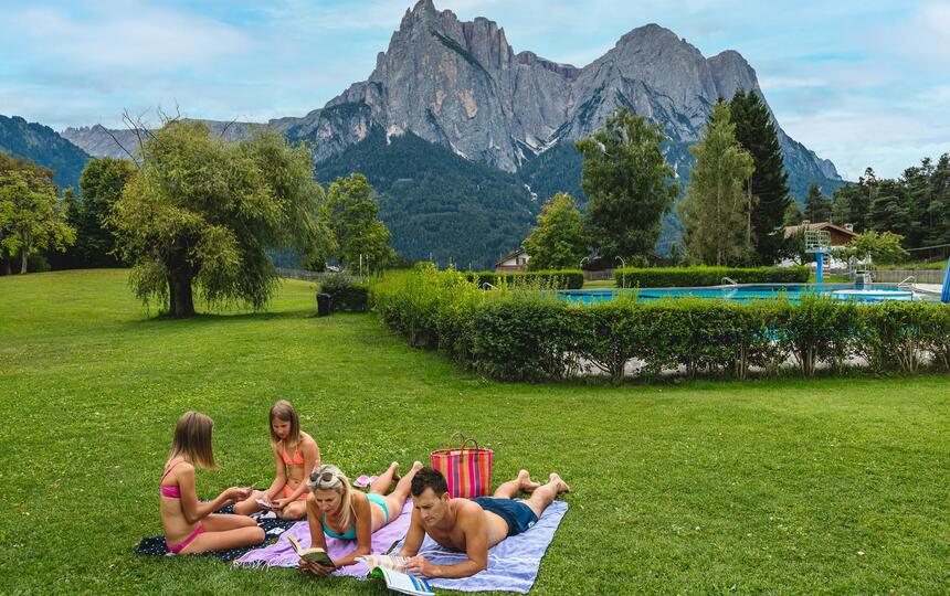 A family is lying on a meadow, with a swimming pool visible in the background
