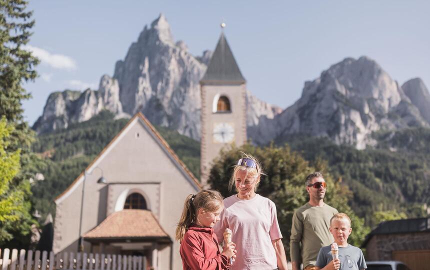 A family with two children is eating ice cream, with a church and the Dolomites in the background