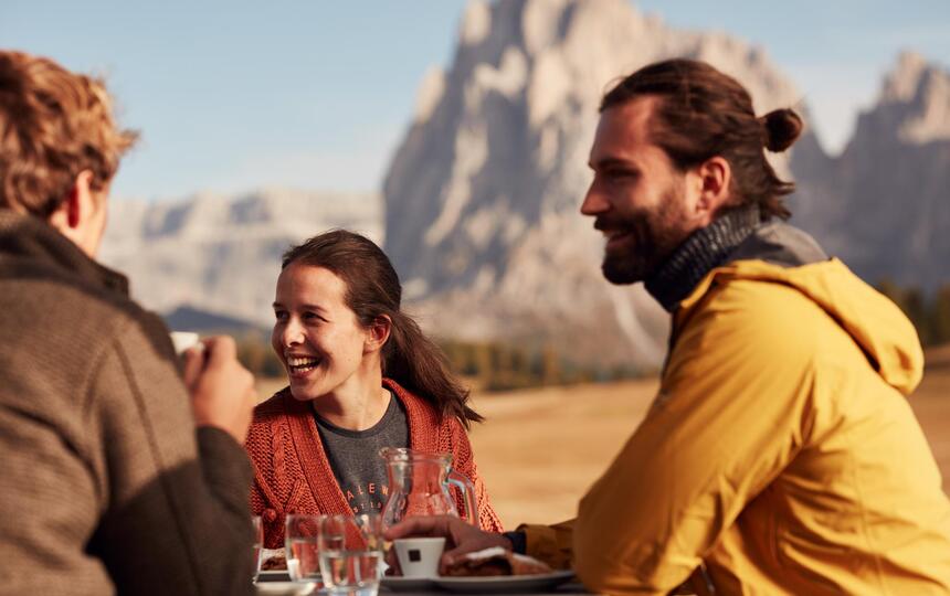 Three people are drinking coffee, with the Dolomites in the background