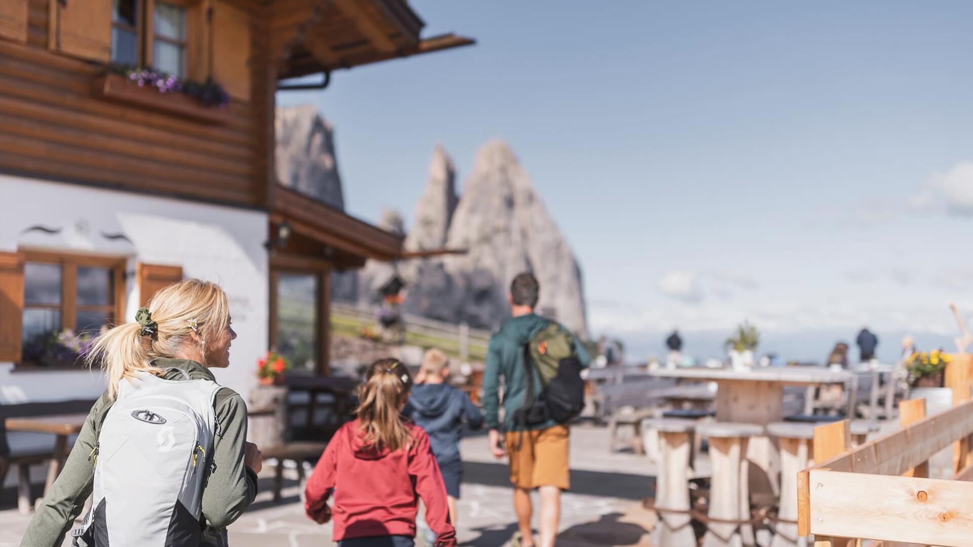 Eine Familie mit zwei Kindern kehrt in einer Hütte ein, im Hintergrund der Schlern
