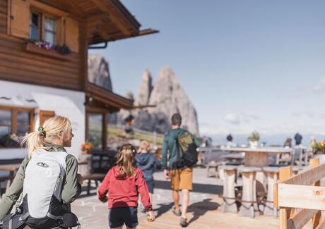 Eine Familie mit zwei Kindern kehrt in einer Hütte ein, im Hintergrund der Schlern