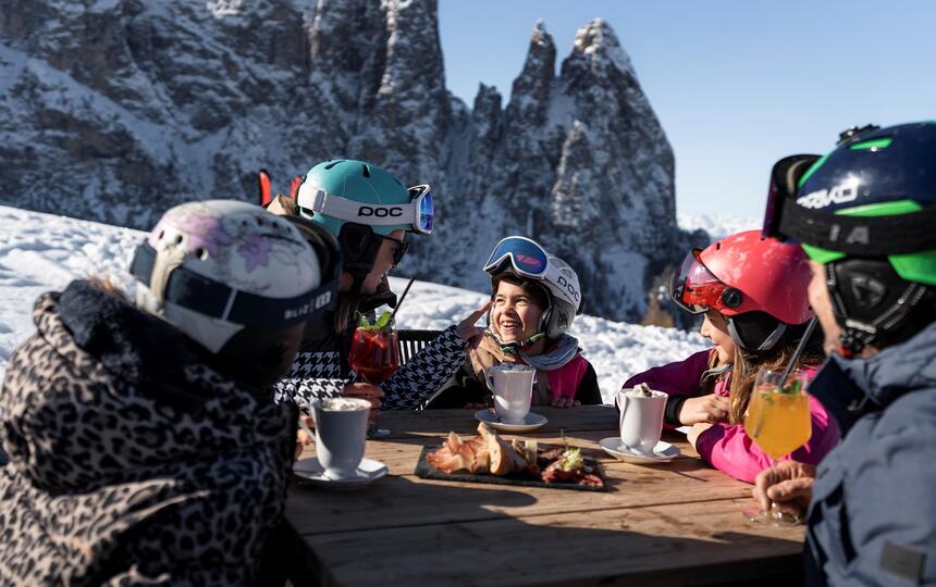 Eine Familie in Skikleidung sitzt im Freien und genießt heiße Schokolade mit dem Schlern im Hintergrund