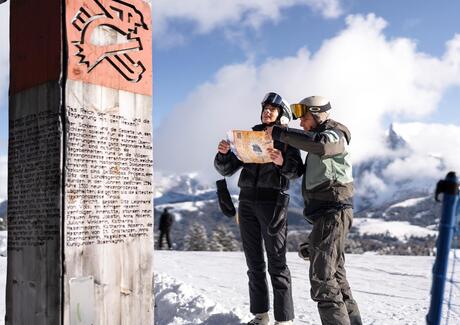 Zwei Personen in Skiausrüstung betrachten gemeinsam eine Karte auf einem Berggipfel neben einem Wegweiser im Schnee