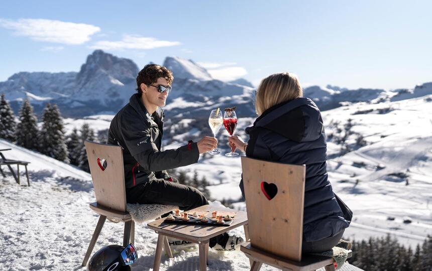 Zwei Personen sitzen im Freien an einem Holztisch, stoßen mit einem Aperitif an und haben Snacks vor sich
