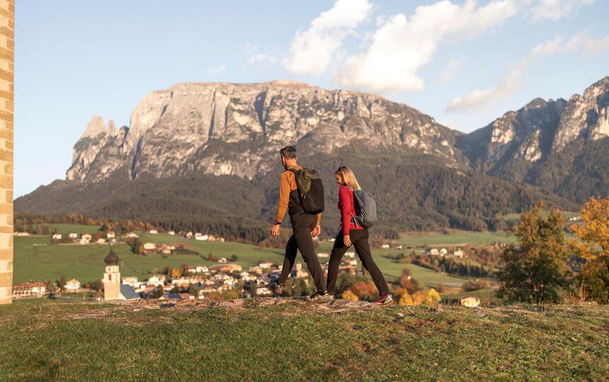 A man and a woman walk along a meadow path and look toward Völs am Schlern