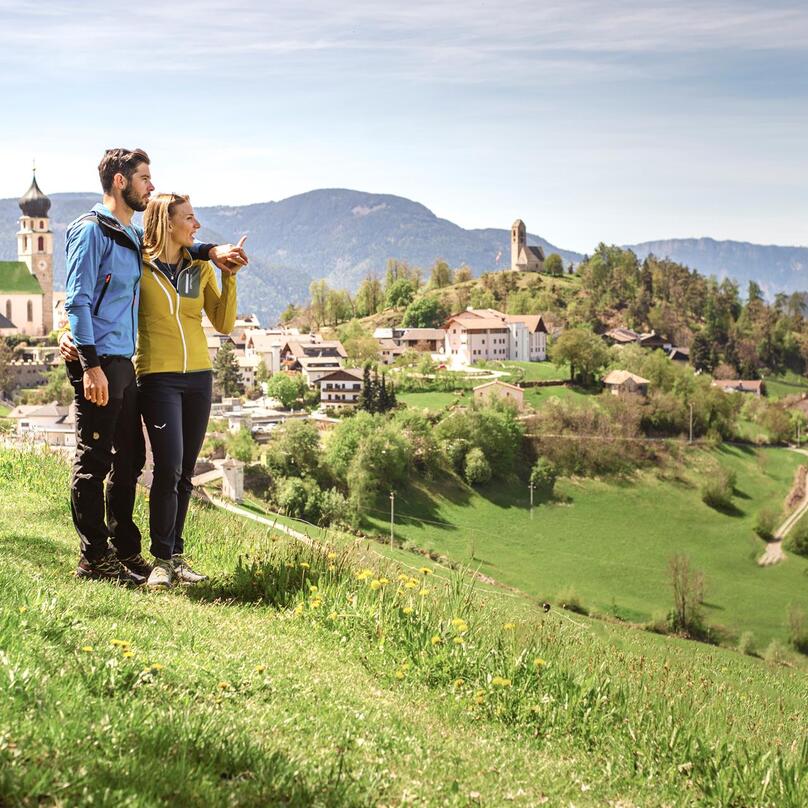 A man and a woman are walking along a meadow; in the background lies Völs am Schlern