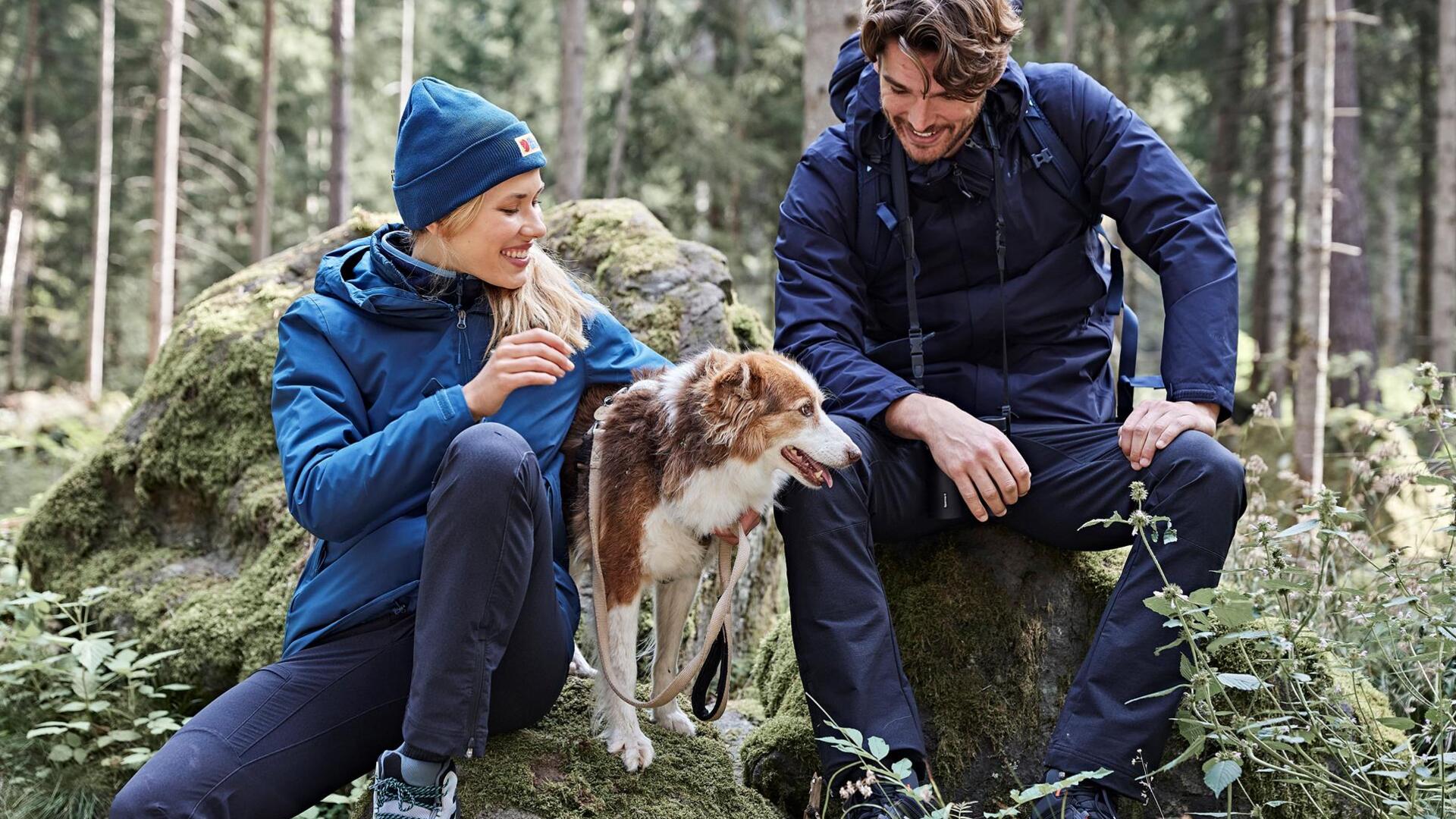 Ein Mann und eine Frau sitzen auf einem Stein im Wald, neben ihnen sitzt ein Hund
