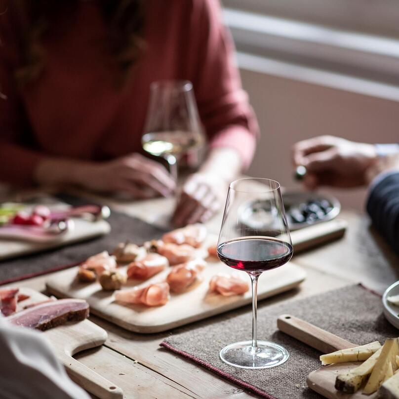 Three people are sitting at a table with a glass of wine, speck and a cheese platter